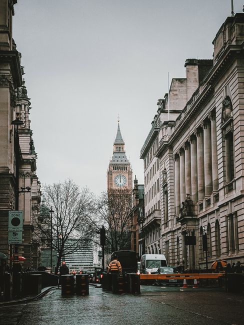 A city street scene in central London showing a narrow road bordered by historic and modern multi-storey buildings with detailed facades. The left side features a large, ornate brick building with dec