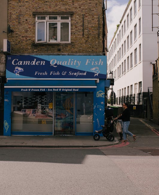 A street-level view of a small fish and seafood shop named 'Camden Quality Fish' with a blue storefront sign; the shop's exterior includes large glass display windows showcasing frozen fish and seafoo