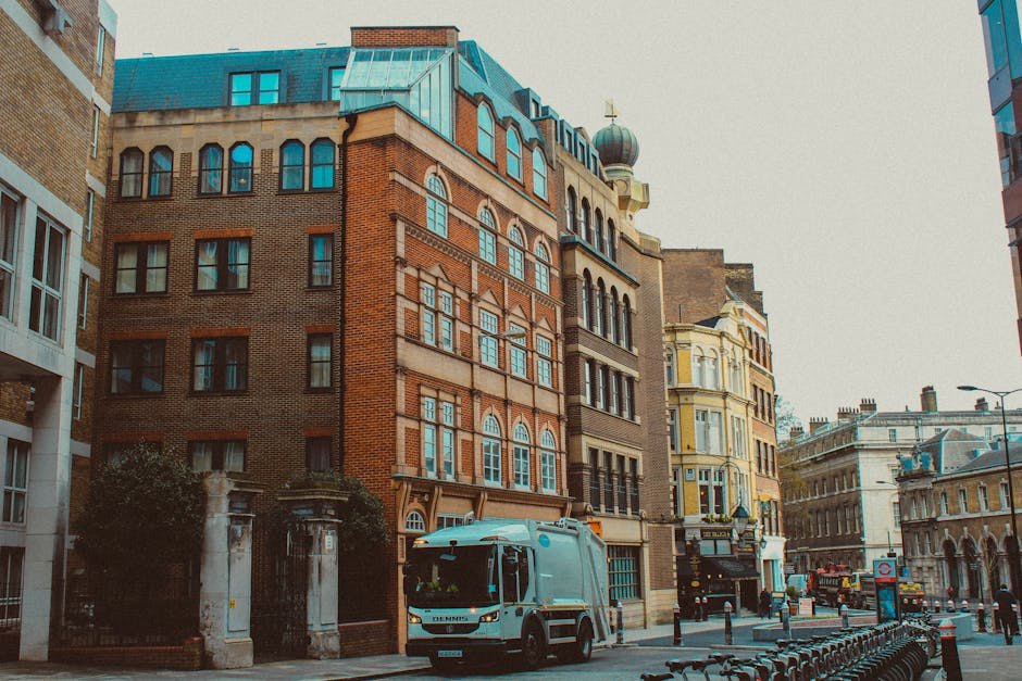 A wet, empty city street scene featuring historic sandstone and brick buildings with ornate window detailing, across from which a row of shops with illuminated signs lines the sidewalk. In the foregro